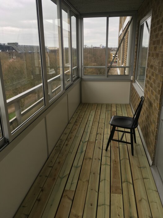 Inglasad balkong med trägolv, stol och utsikt över park.
