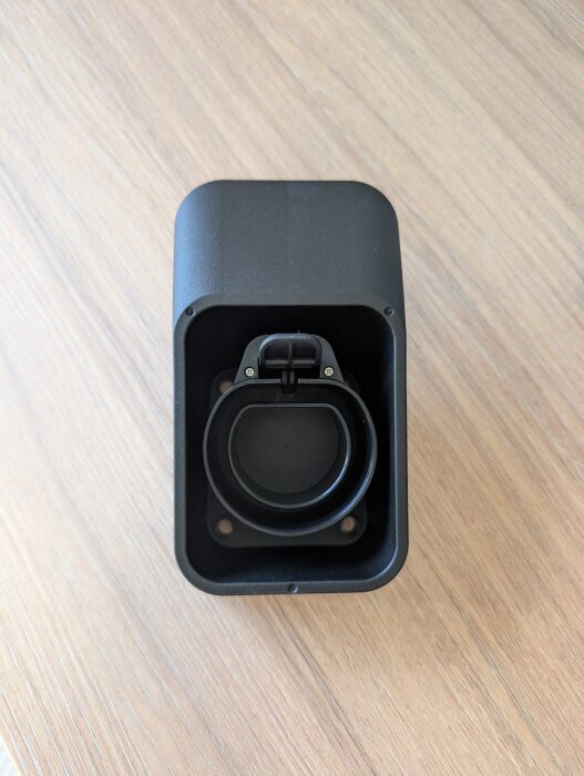 Svart kabelhållare för laddstolpe, liggandes på träbord, med fyra synliga skruvhål.