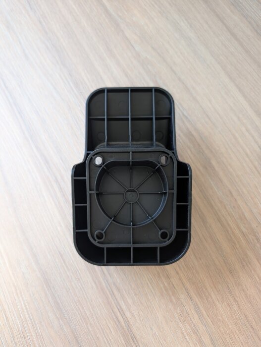 Svart kabelhållare med rutmönstrad baksida och synliga skruvhål, ligger på ett ljust trägolv.