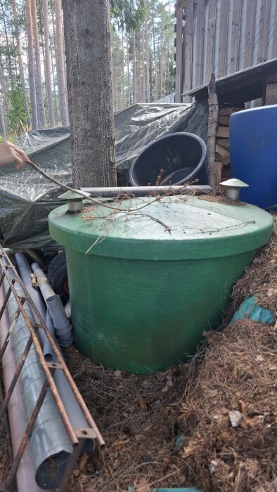 Stor grön, rund behållare med två ventilationer på toppen, placerad på en tomt bredvid en trädstam, metallrör och bråte.