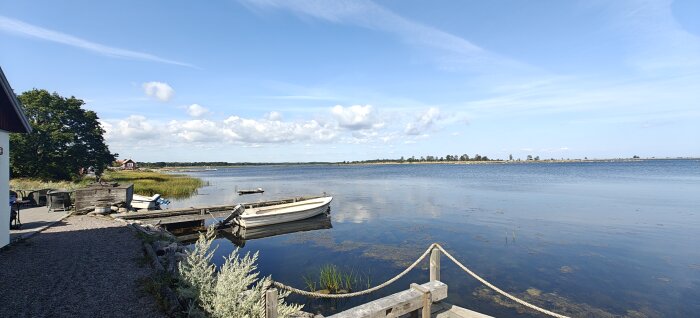 Utsikt över en lugn sjö med blå himmel, en båt vid bryggan, grönska och en stuga i bakgrunden.