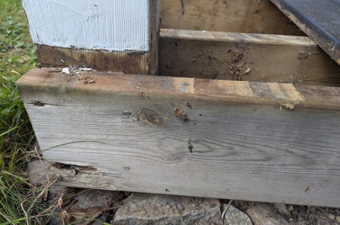 Sliten träregel och stolpe med skadad och avskalad färg vid balkong, omgiven av gräs och stenar.