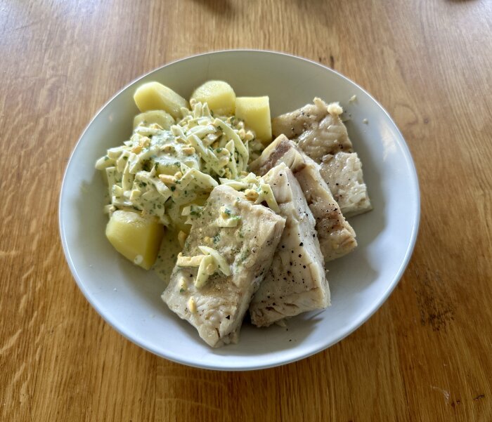 En vit tallrik med kokt fisk, skivad potatis och krämig fyllning dekorerad med örter, på ett träbord.