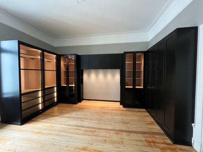 Exklusiv garderob och förvaringslösning med svarta skåp, glasdörrar, träinredning, LED-ljuslister och gott om förvaring, fotograferad i ett ljust rum.