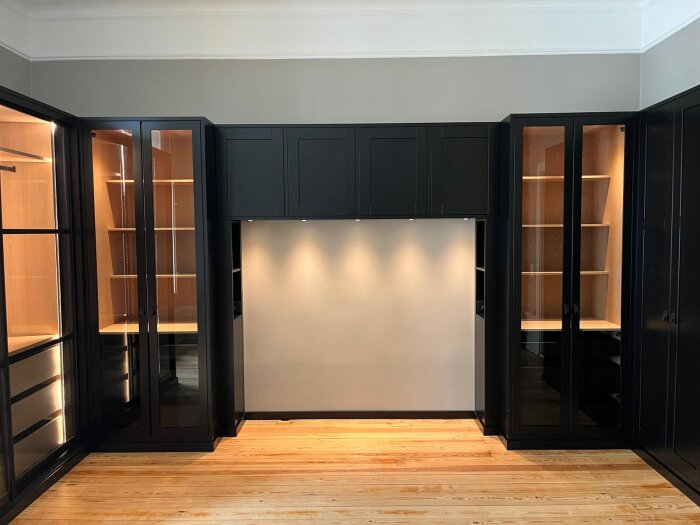 Exklusiv garderobsinredning med svarta glasdörrar, träinredning och inbyggd LED-belysning, mot en ljus vägg och trägolv.