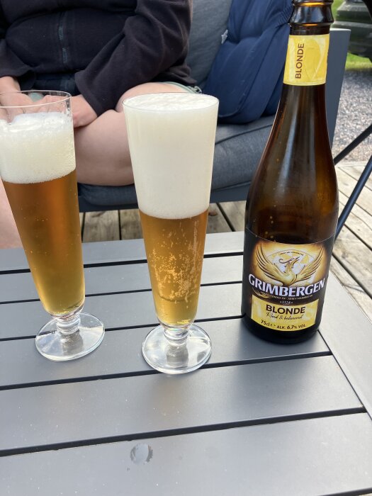 Två höga ölglas med skummande öl bredvid en flaska Grimbergen Blonde på ett bord utomhus. En person sitter i bakgrunden.