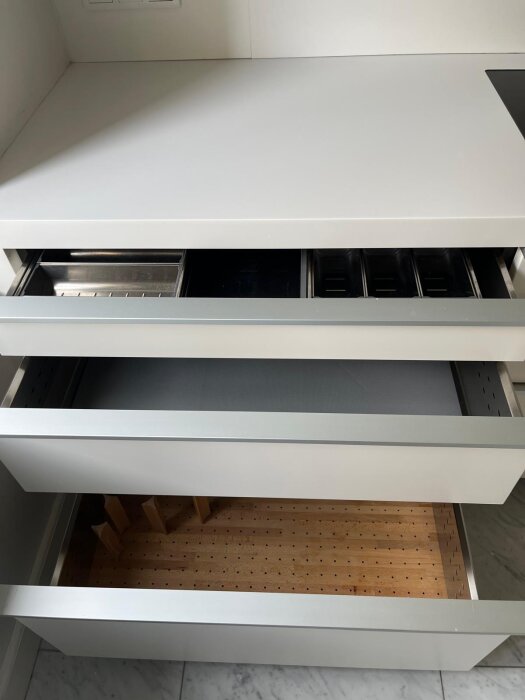 Tre öppna kökslådor från Minotti med olika inredning, inklusive bestickinsats i metall och trälåda för tallrikar, i ett kök med varmvita ytor.