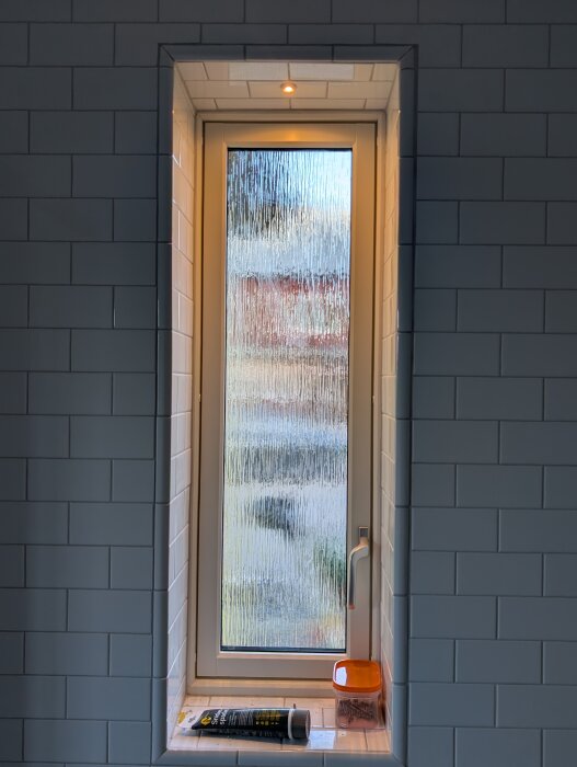 Liten lampa monterad i väggen ovanför ett smalt fönster med texturerat glas, omgiven av vit kakelvägg.