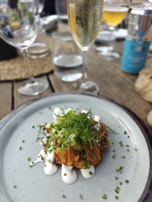 Kantarelltoast med grönsaker och dill på en tallrik, omgiven av vinglas och en blå dryckesburk på träbord.