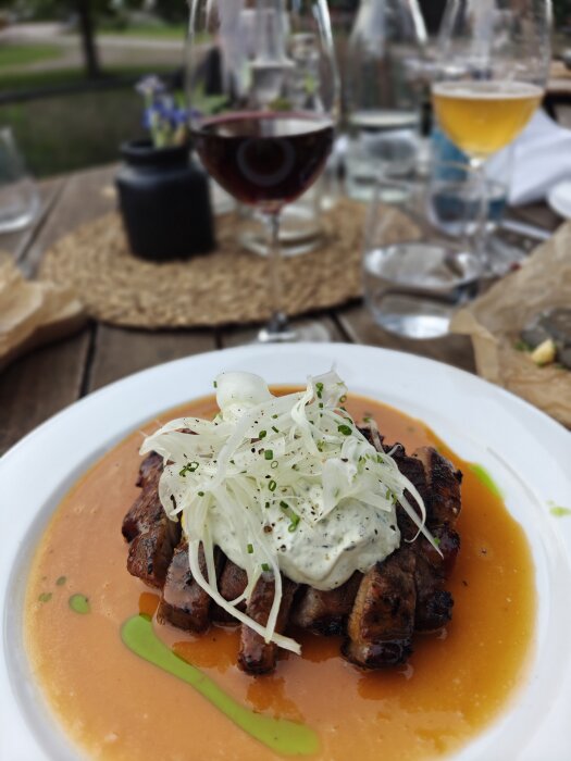 Tallrik med köttskivor, toppade med sallad och sås, i en orange sås. Ett glas rött vin i bakgrunden på en träbord.