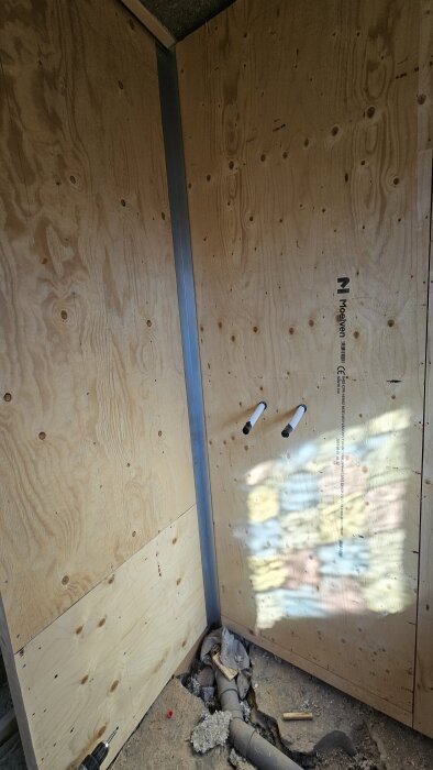 Vägg av plywoodpaneler och gipsskruvar, med utskjutande metallrör. Golvet i trä och betong med rör och bråte.