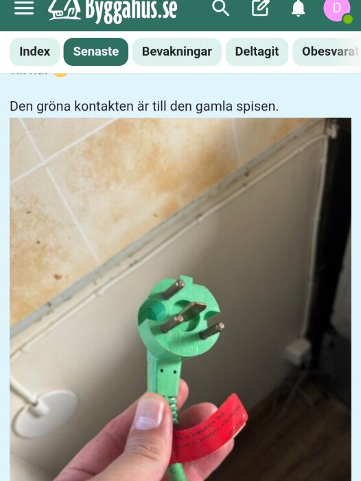 Grön elkontakt till en spis, hålls i en hand, med röd varningsflagga i bakgrunden av ett kök.