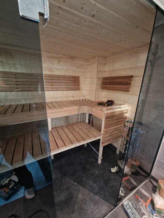 Bilden visar en bastu under renovering med träsitsar och glasdörr, verktyg och material syns på golvet.