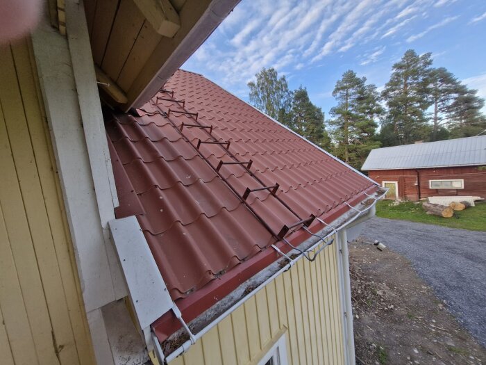 Rött plåttak med en demonterad hängränna sedd från sidan av ett gult hus, omgiven av skog och en liten väg.