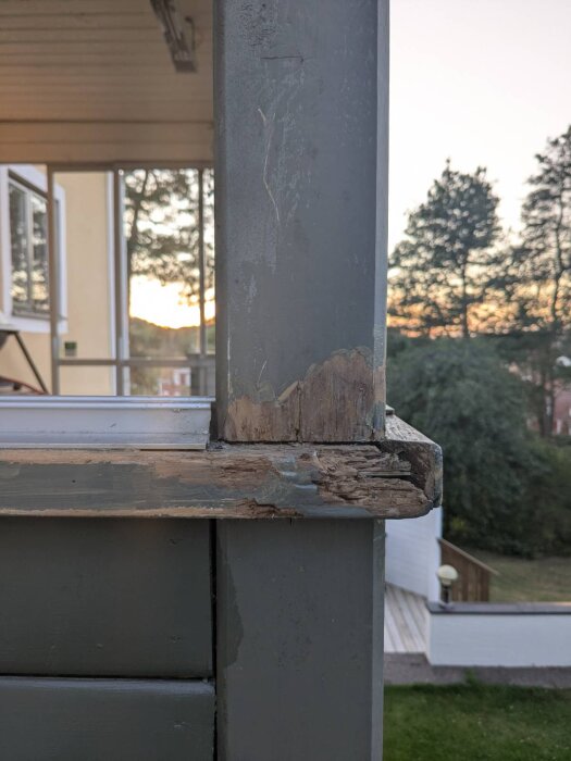 Närbild av skadad balkongstolpe och plankor med flagnande färg och träskador. Stolpen sträcker sig upp mot taket. Skog och himmel i bakgrunden.