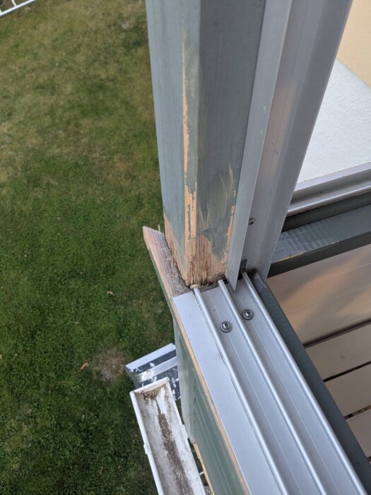 Skadad trästolpe på balkong med avslitna och nötta partier, metallprofiler och vy mot gräsmatta i bakgrunden.