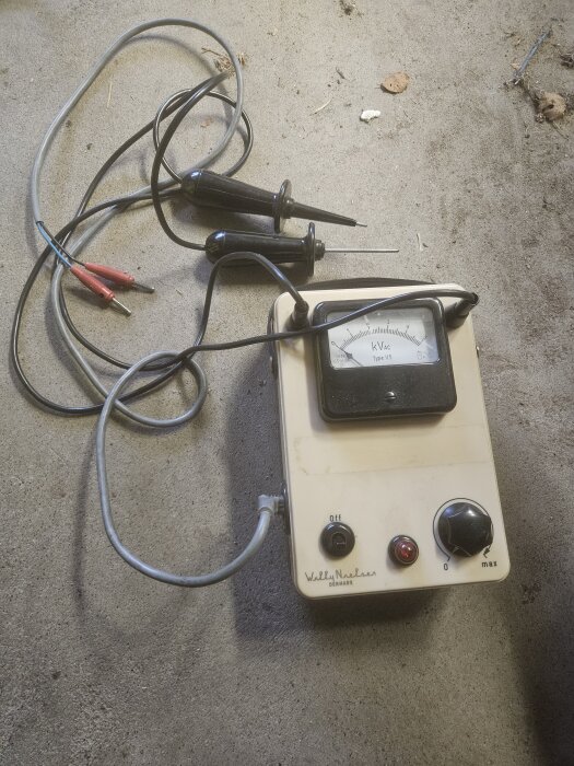 En vit enhet med en kvadratisk mätare märkt "kV AC", två testprober och en vridknapp. Texten säger "Wally Neaders, Denmark".