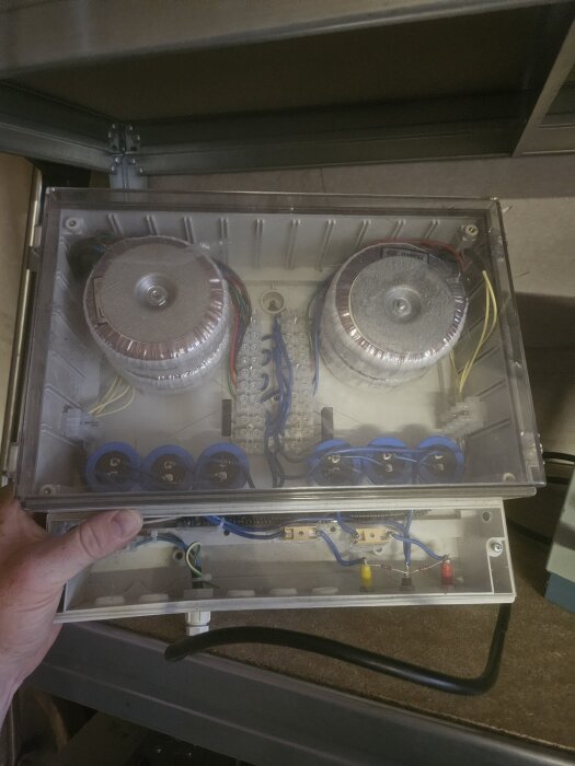Box med två transformatorer och komponenter för likriktning av 230VAC till 230VDC, synliga ledningar och kontakter, hålls i handen.