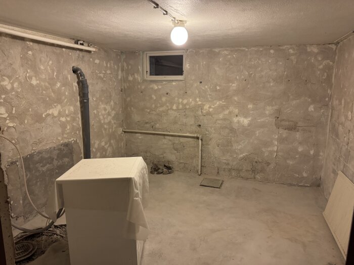 Källarrum under renovering med synliga cementväggar, ett ensamt rör, ett stuprör och ett golv där det börjat bilas runt rören. En lampa hänger i taket.