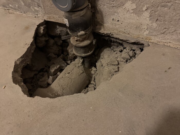 Hål i betonggolv i källare med synligt trasigt gjutjärns- och betongrör samt smuligt material runtomkring.