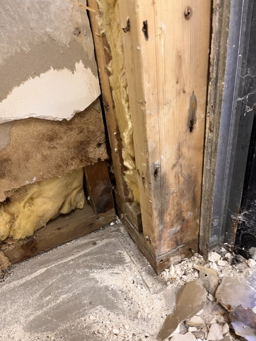 NÀrbild av ett garagehörn med synlig trÀregel, isolering och sprucken vÀggputs. Det finns spÄr av fukt och damm pÄ betonggolvet.