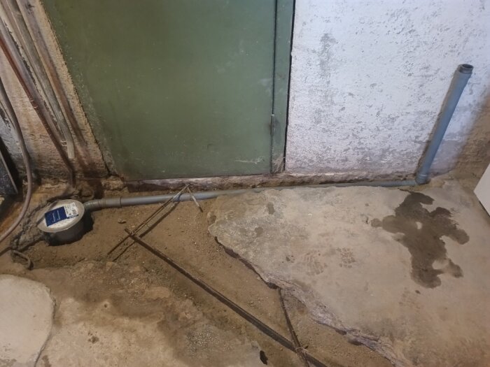 Rörinstallation i källare före gjutning, med framträdande avloppssystem och rostfri brunn vid betonggolv och dörr.