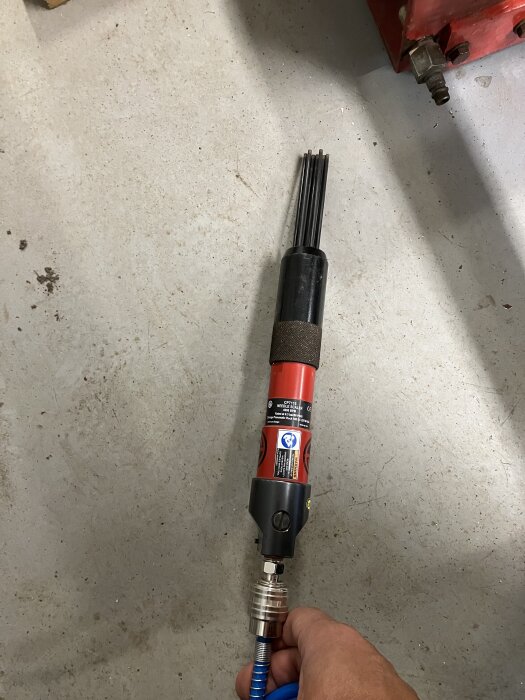 Pneumatiskt verktyg med nålhammare används för att ta bort färg och rost från ett stålräcke inför målning, hand håller i verktyget.