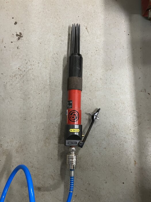 Pneumatisk nålhacka med röd och svart design, blå luftslang ansluten, använd för att avlägsna färg och rost från stålräcke.