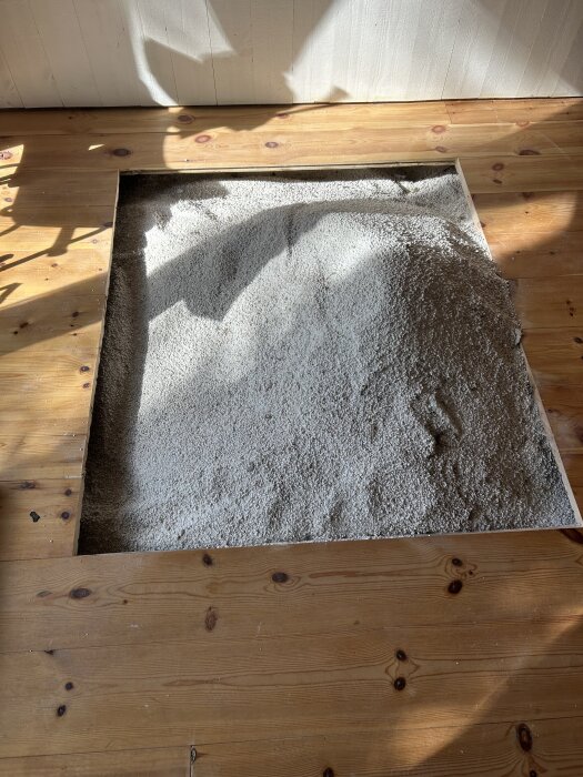 Öppet golvparti med synlig perlite under trägolv i oventilerad torpargrund, skuggor och ljus spelar över ytan.