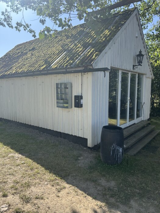 En vit byggnad med mossbelagt tak, glasdörrar och en svart tunna vid trädgården under ett träd i solsken.
