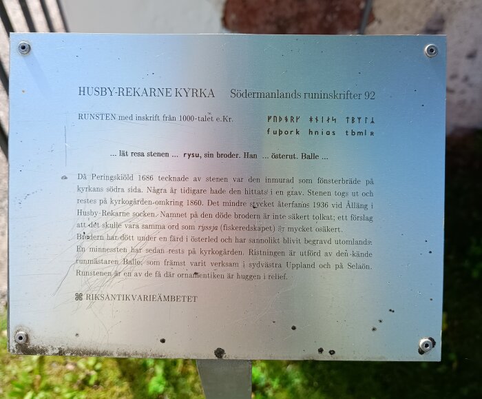 Informationsskylt om runstenen vid Husby Rekarne kyrka. Text om ristningar av runmästaren Balle, ursprung och historik. Nummer: Södermanlands runinskrifter 92.