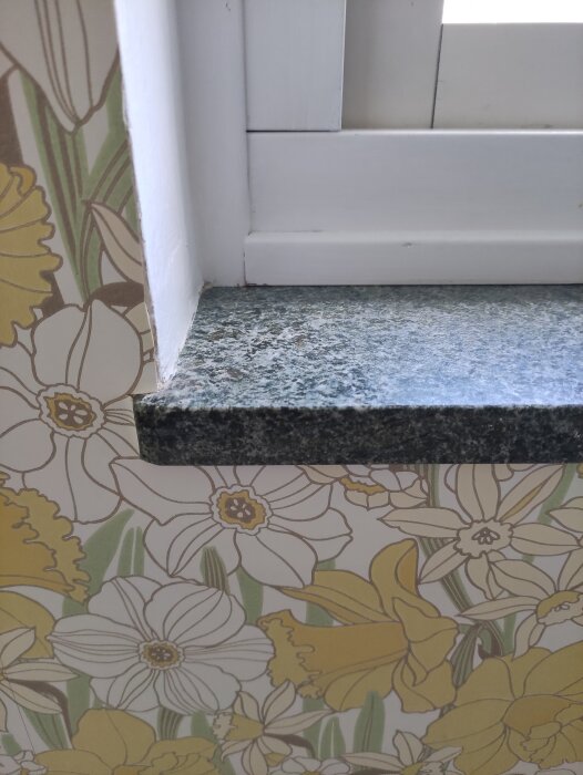 Fönsterbänk i mörkgrön sten vid fönster med färgglad blomstertapet i bakgrunden.