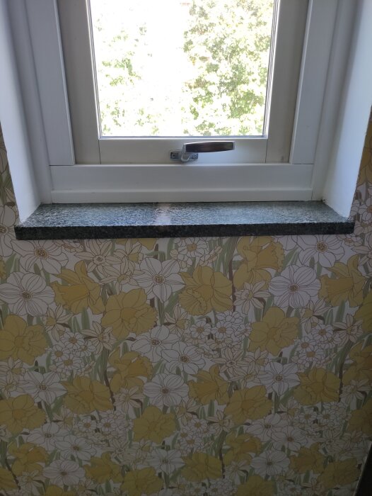 Fönsterbänk i mörk sten, möjligtvis Verde Savana granit, med blommigt tapetmönster i gult och vitt i bakgrunden.