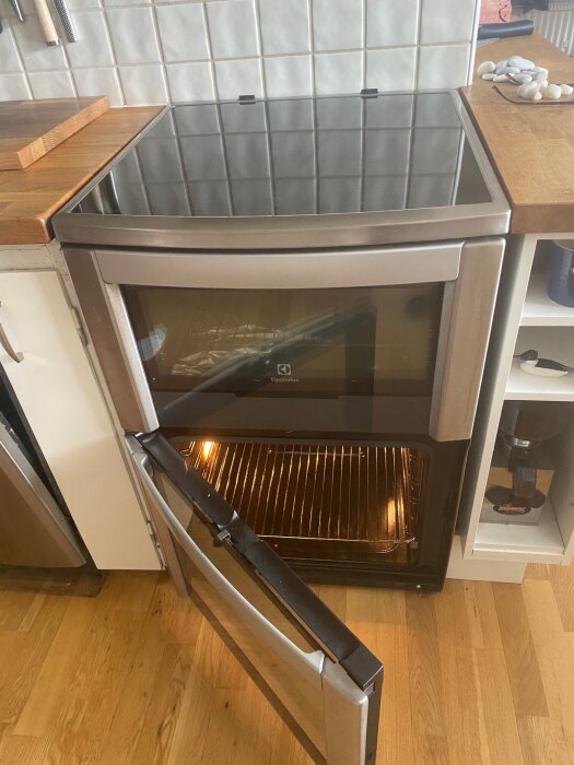 Spis med öppen ugnslucka, keramikhäll ovanpå, placerad i kök med vit kakelvägg.