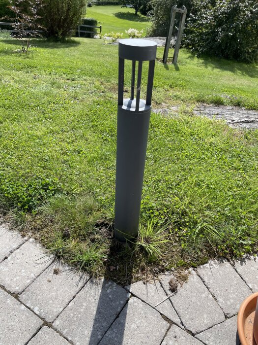Grå utomhuslampa på gräsmatta invid stenplattor i solsken, del av utebelysning i tråddiskussion om JFB-problem.