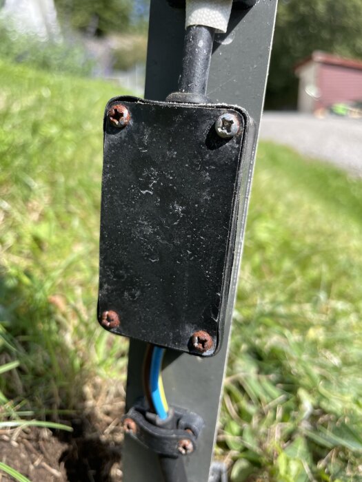 Transformatorlåda monterad på en stolpe med synliga kablar och rostiga skruvar i en trädgårdsmiljö.