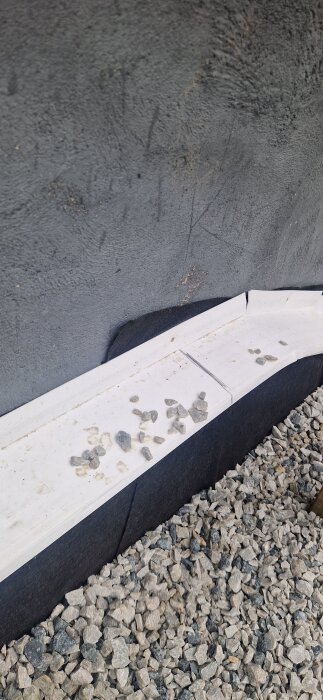 Grus längs husgrundens vägg på en sluttningstomt, dräneringslist och markduk synliga, några småstenar ovanpå listen.