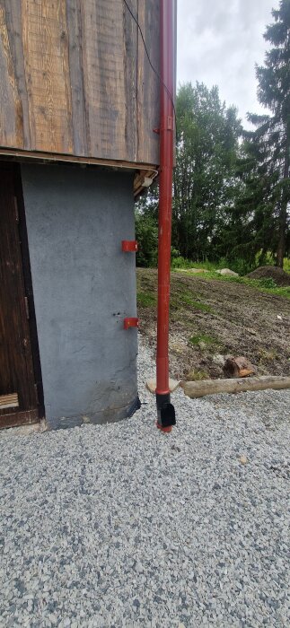 Ett hus fasad med träpanel, synligt stuprör och grusbädd framför. Grå vägg och röd stuprör. Träd i bakgrunden.