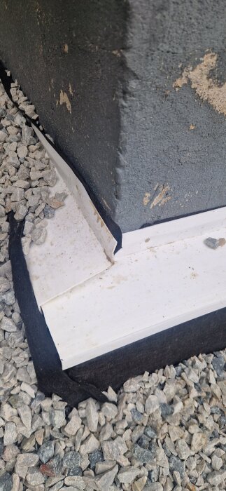 Grund med isolering och grus runt ett hörn av ett hus. Isoleringen går ovan marken och är omgiven av markduk och grus.