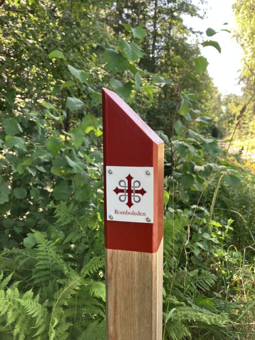 En träpåle med rött topp och skylt med texten "Romboleden" står omgiven av grönska i skogsmiljö.