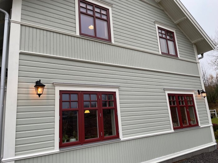 Fasad av ett hus i äldre stil med moderna fönster och utomhusbelysning, vitmålat trä och röda karmar.