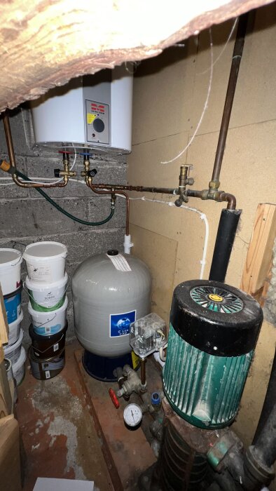 Äldre vattenpump och hydropress i källare, omgiven av rör och färgburkar. Manometer och ventil synliga bredvid pumpen.