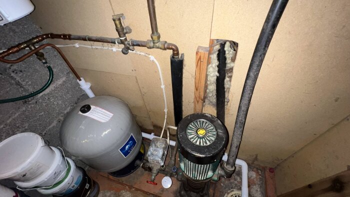 Äldre vattenpump och hydropress i ett källarutrymme med rörsystem, mot en vägg.