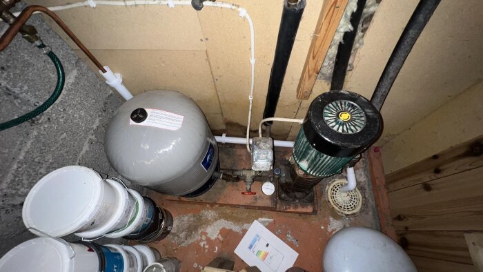 Vattenpump och hydropress i äldre hus med tillhörande rörledningar och tryckmätare, belägen i källarutrymme med målad betonggolv.