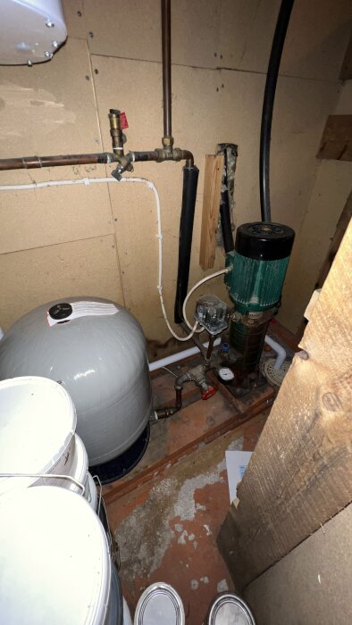 Äldre vattenpump och hydropress installerade i källare med olika rörkopplingar, tryckmätare och ventiler synliga på väggen.