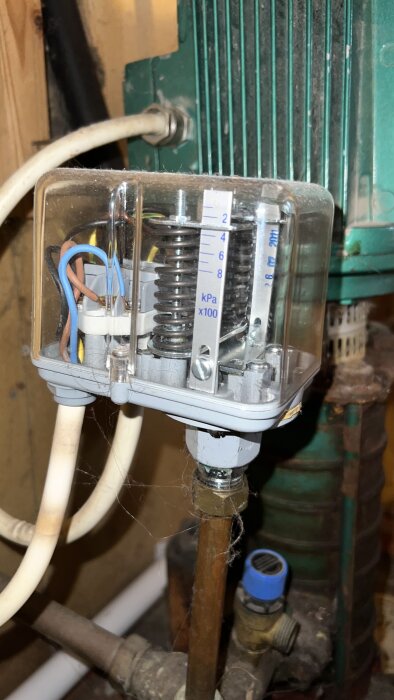 Närbild på en tryckströmbrytare med synliga fjädrar och kablar, monterad på ett vattenpumpningssystem, delvis täckt av damm och spindelväv.