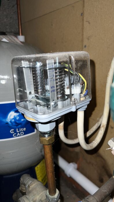 Närbild på tryckströmbrytare med synliga fjädrar och elkablar, monterad på kopparrör, del av vattenpompsystem i äldre fastighet.