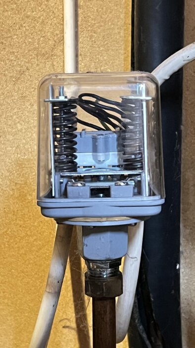 Tryckströmbrytare monterad på ett vattenrör med flera kablar och fjädrar synliga inuti ett transparent plasthölje.