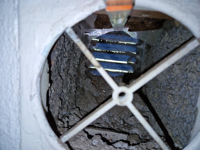 Bilden visar en närbild av ett ventilationsschakt med synliga sprickor och ett metallgaller.