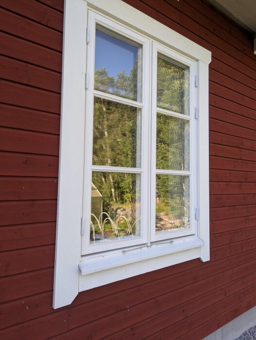 Rött trähus med ett vitt fönster. Fönstret reflekterar omgivande träd och blå himmel.